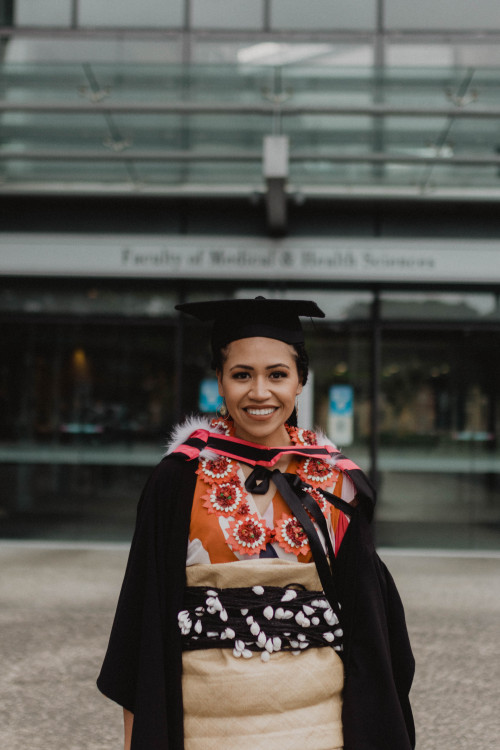 waist upwards photo of Dr Paea Leakehe dressed in traditional clothing and graduation gown standing in front of Faculty of Medicine and Science Builidng 